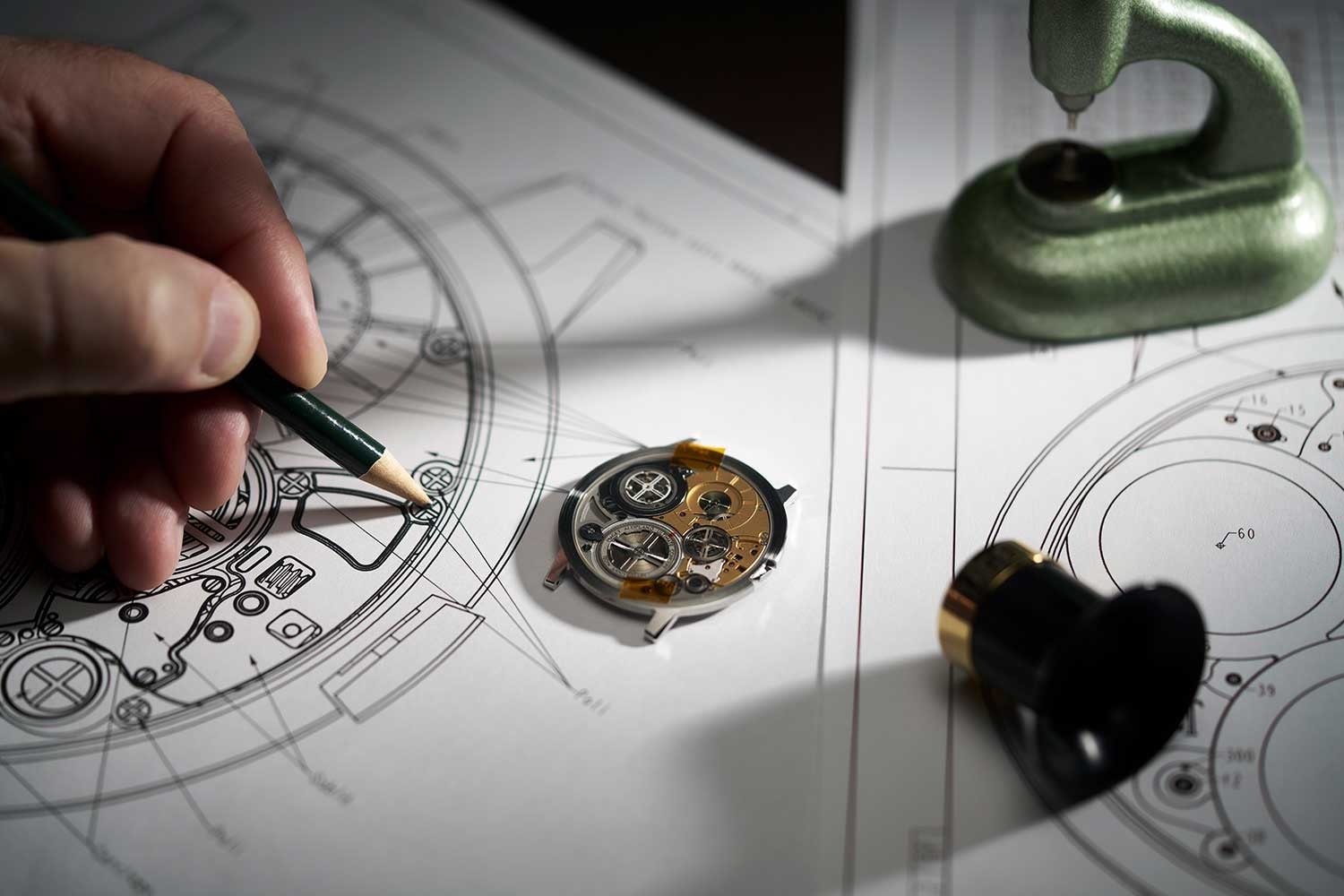 Hand sketching a custom watch over a technical blueprint beside a partly assembled movement